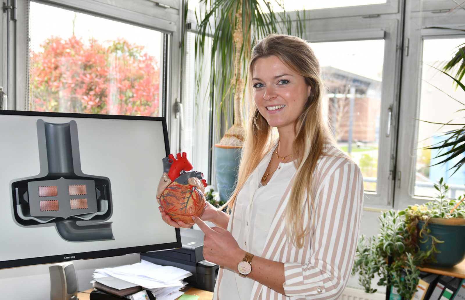 Theresa Gärtner mit einem Modell des menschlichen Herzens. Auf dem Bildschirm ist das Implantat zu sehen mit der wellenförmigen Bewegung der Membran. (Foto: FH Münster/Katharina Kipp) 