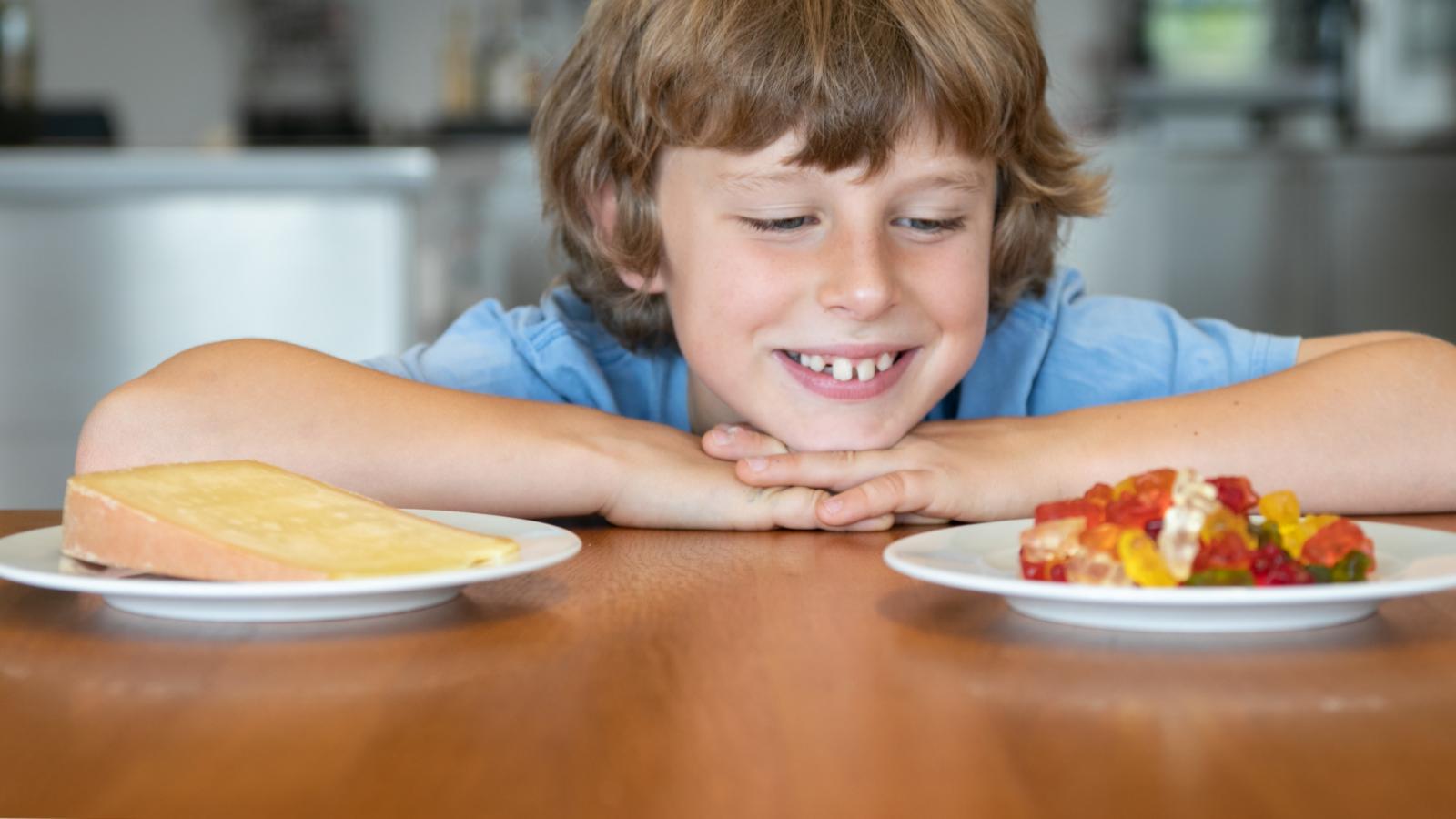Ein Kind sieht zu einem Teller Gummibärchen auf einem Tisch. Daneben steht ein Teller mit Käse.