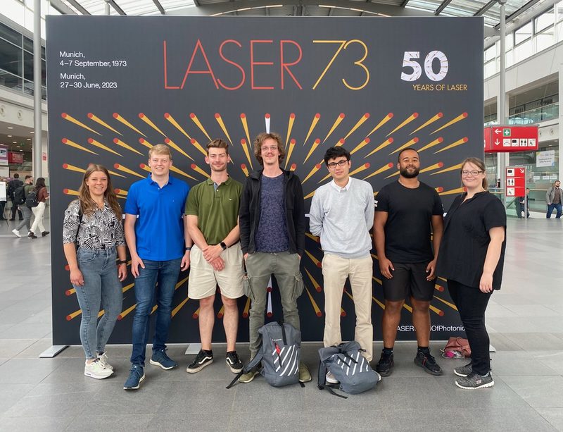 Eine Gruppe von sieben Studierenden posiert vor einem großen Plakat der Lasermesse